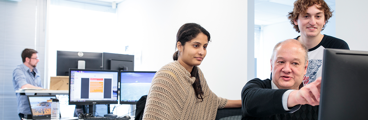 Two people standing around a man on a computer pointing at his screen